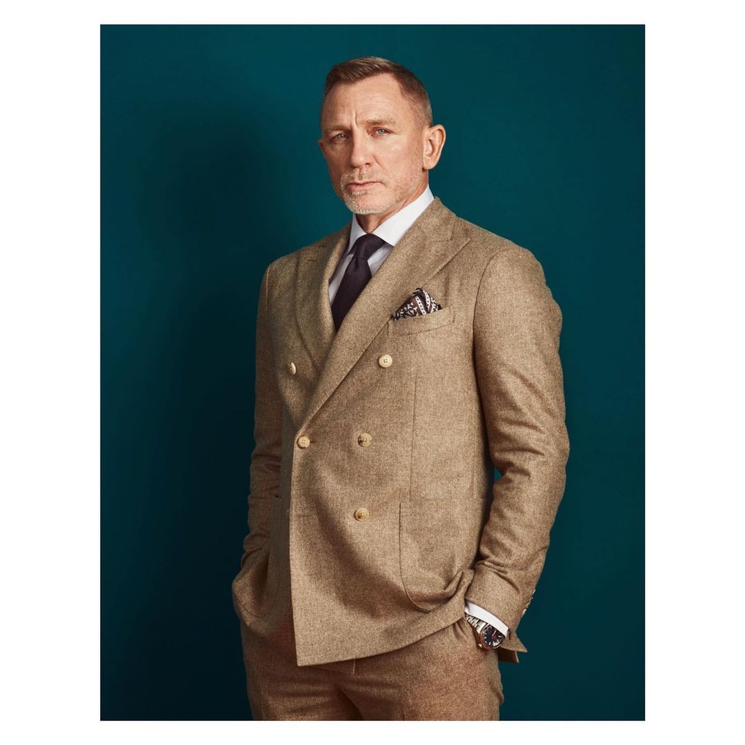 Portrait of an adult male wearing a tailored brown suit, black tie, and patterned pocket square, set against a solid teal backdrop.