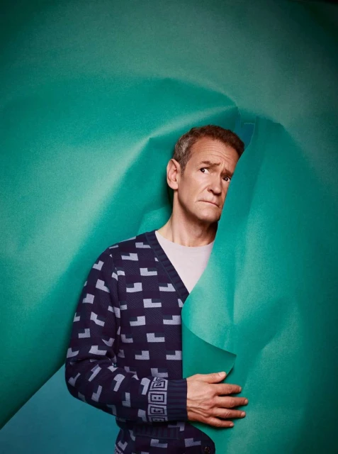 An adult male poses against a teal paper background while wearing a patterned cardigan, looking to the side with a slight frown.