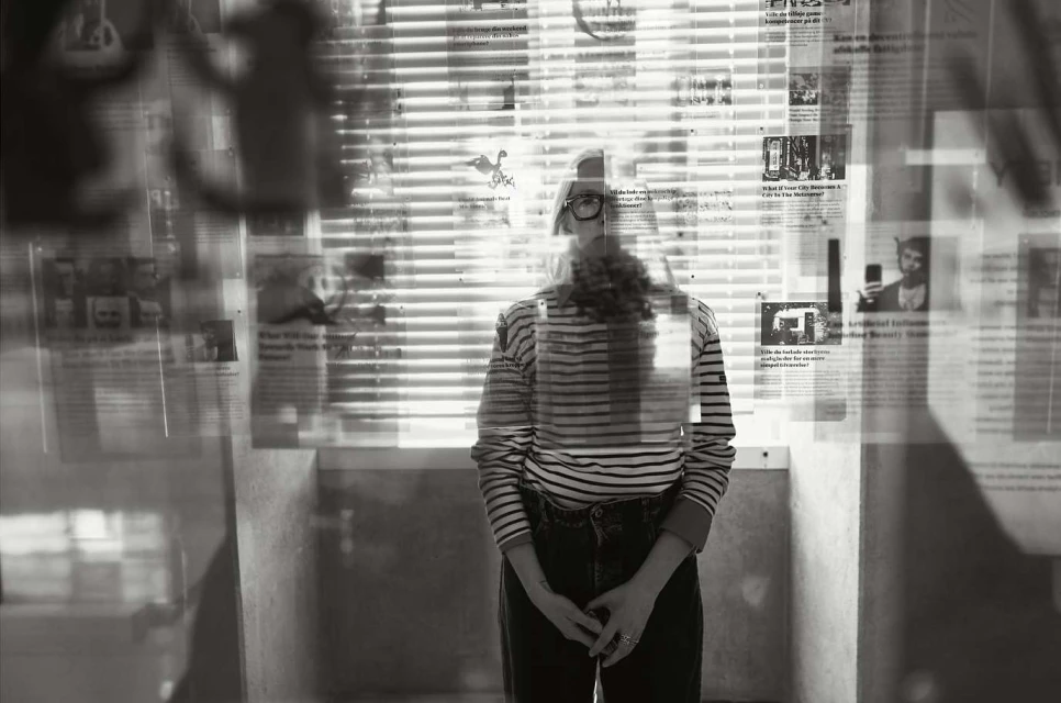 Black-and-white portrait of a woman with glasses, wearing a striped shirt, posing in front of a window with newspaper clippings.