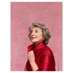 Elderly woman with short grey hair wearing a high-collared red blouse against a textured pink background.