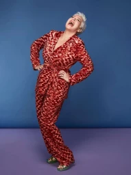 Laughing older woman with white hair wearing a patterned red velvet pantsuit against a blue backdrop.