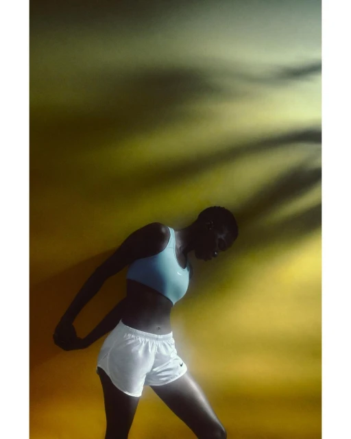 Female athlete stretches against a yellow background with tree shadow. Woman wears a sports bra and running shorts in a fitness setting.