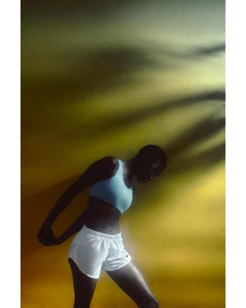 Female athlete stretches against a yellow background with tree shadow. Woman wears a sports bra and running shorts in a fitness setting.