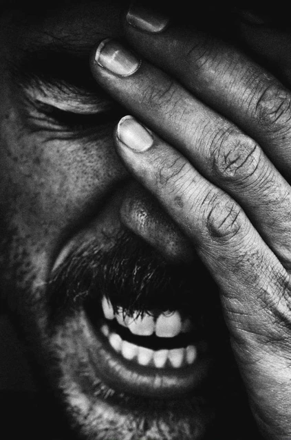 Close-up black and white portrait of an adult male with hand covering part of his face, revealing a wide smile and expressive features.