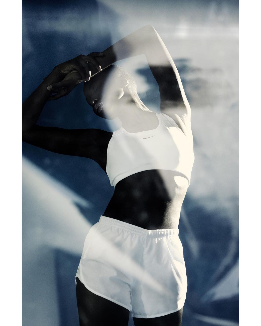 Athletic woman in white sports bra and shorts stretches her arms overhead against a blue background with diffused light and shadows.
