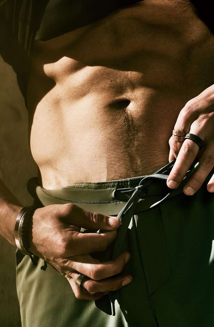 Athletic male torso with defined abs and olive athletic pants, adjusting drawstring, with a background of a neutral, textured wall.