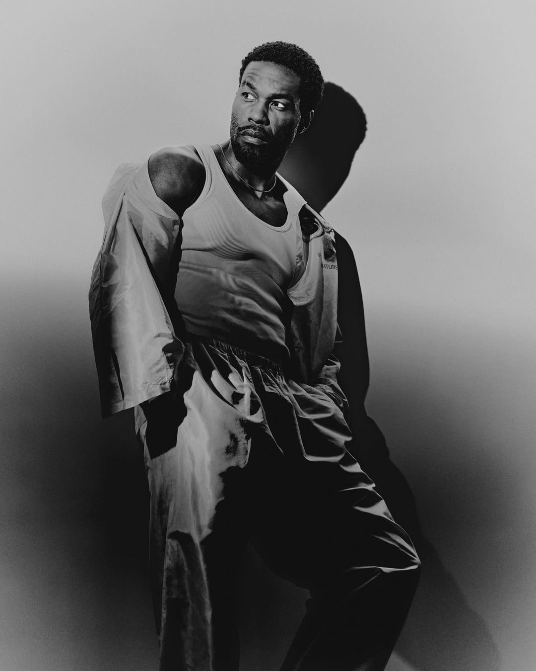 Black and white portrait of an adult man in casual clothing against a grey backdrop with visible shadow detail, looking off-camera.