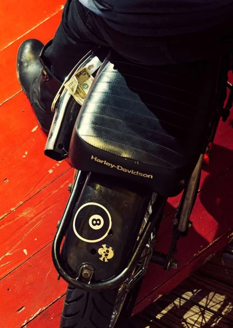 Closeup of a person sitting on a Harley-Davidson motorcycle with a black leather seat, an 8-ball sticker, and money attached.