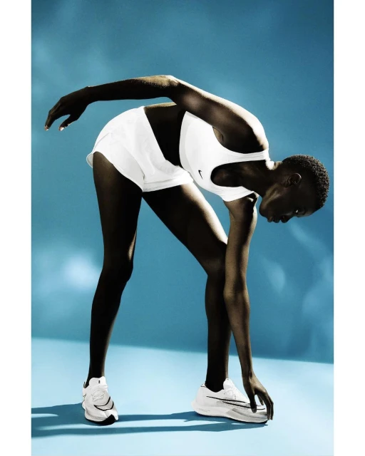 Athletic African American woman in white sports bra and shorts bends over stretching her leg, wearing running shoes against a blue backdrop.