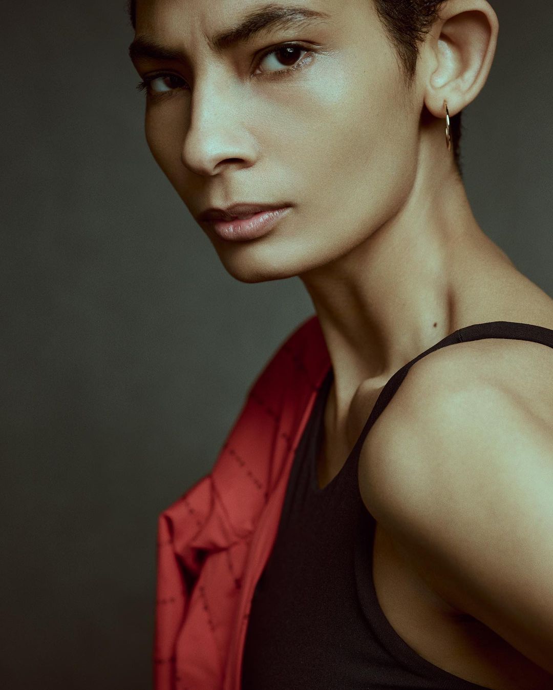 Portrait of a young adult with a short hairstyle, wearing a black top and a red jacket over her shoulder, posing against a grey background.