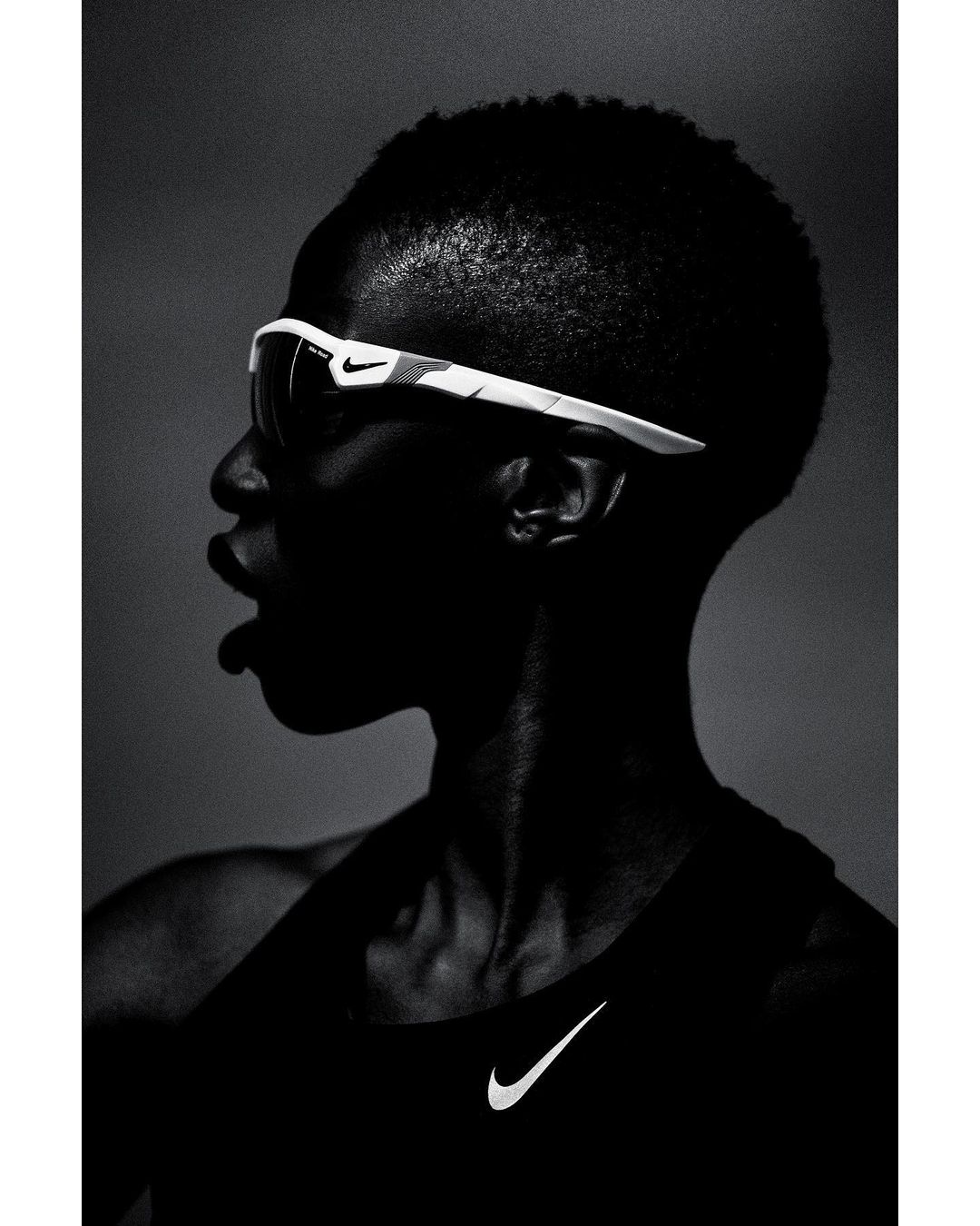 Profile shot of a dark-skinned adult wearing sunglasses and a tank top with a Nike logo against a dark grey gradient background.