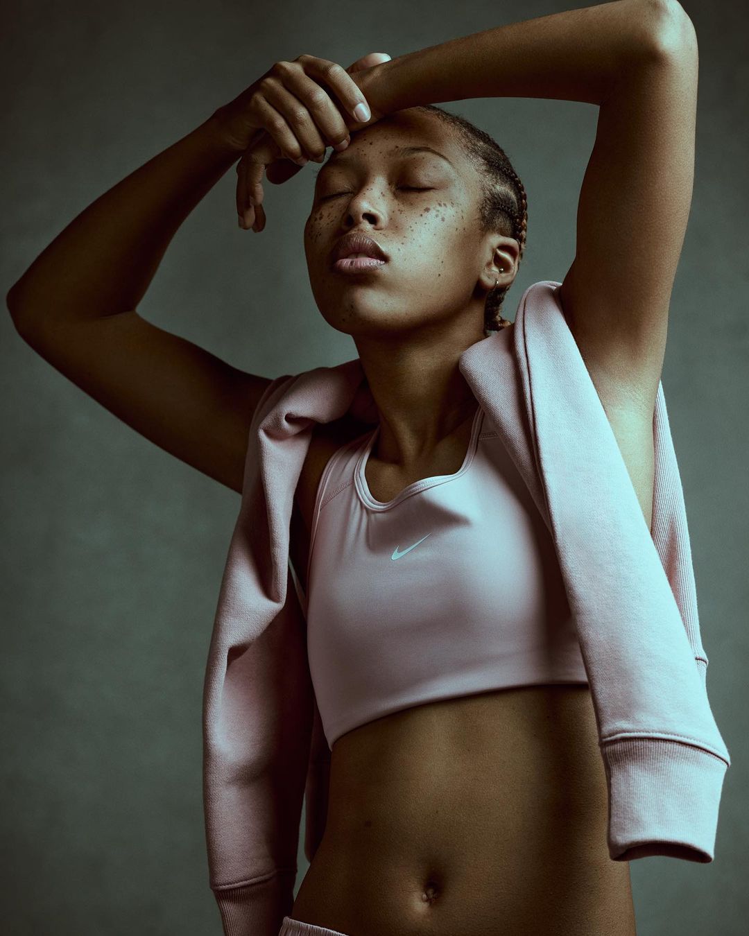 Athletic woman in pink sports bra and track suit poses with hands above head against a muted grey backdrop in a studio shot.