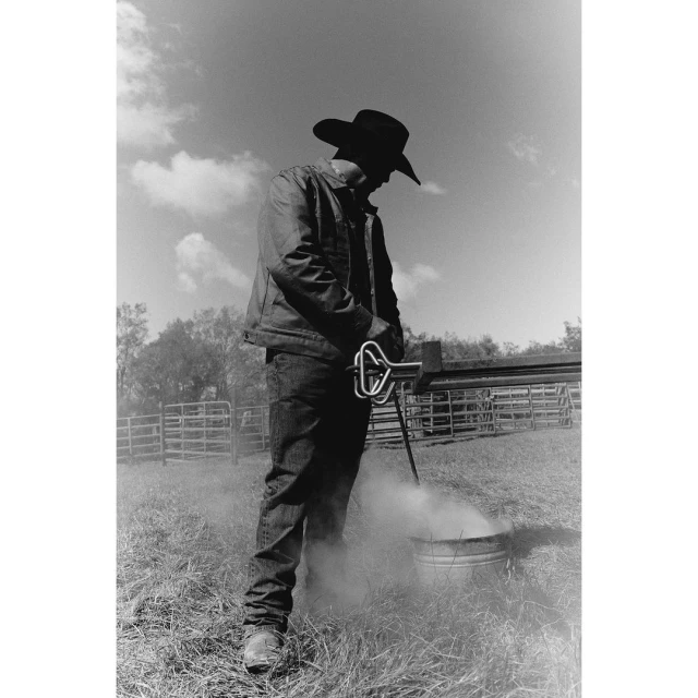 Monochrome photo of a silhouetted cowboy in hat and jacket tending to a metal bucket with branding iron and steam rising in a grassy field.