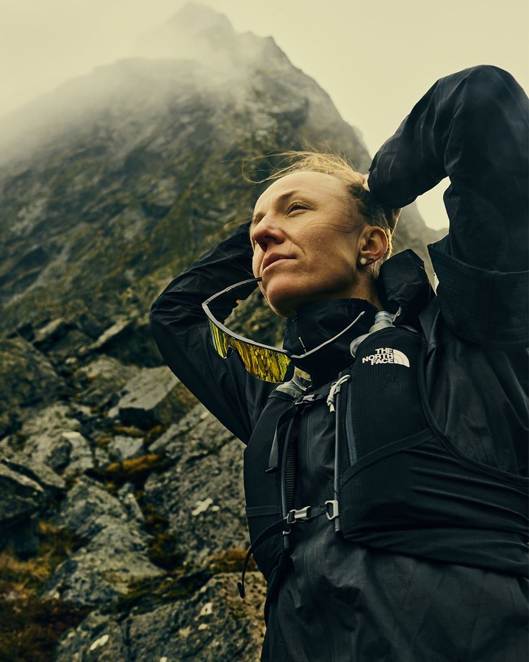 Adult female trail runner pauses on rocky mountain, hands on head, wearing black jacket, hydration pack, sunglasses, outdoor setting.