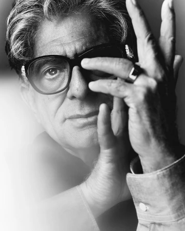 Close-up monochrome portrait of an older man wearing dark oversized glasses and a ring, with a hand raised near his face.