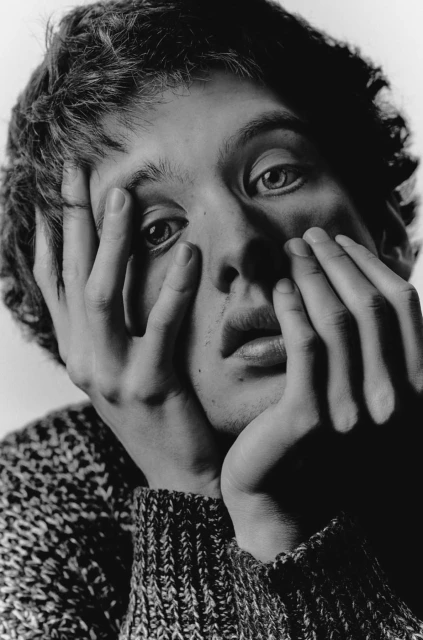 Monochromatic portrait of a young adult with hands on their face, wearing a textured sweater against a white backdrop.