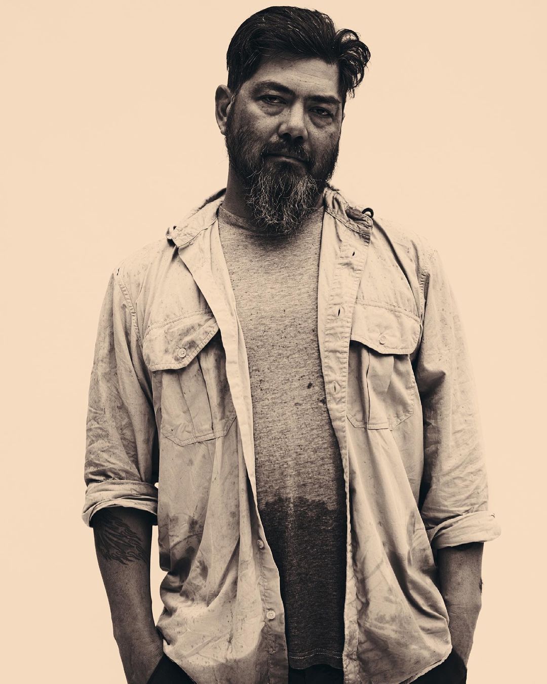 Portrait of a bearded man with an open collared shirt, gazing directly into the camera with a neutral expression in a studio shot.