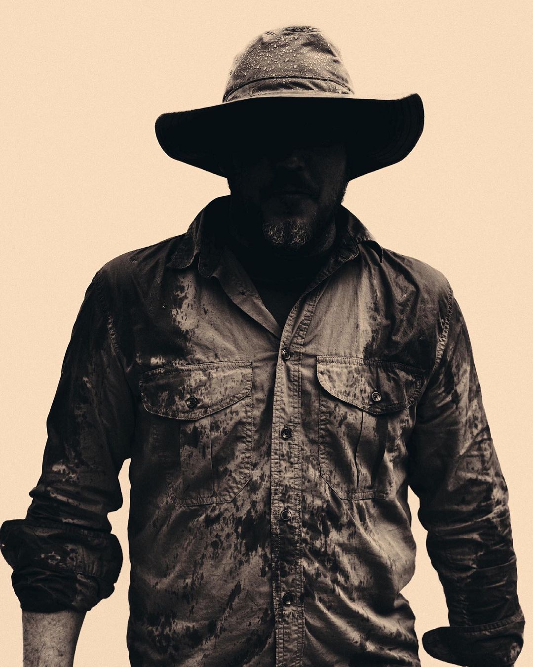 Monochrome portrait of a bearded adult male wearing a hat and buttoned work shirt with spots covering the garment against a tan backdrop.