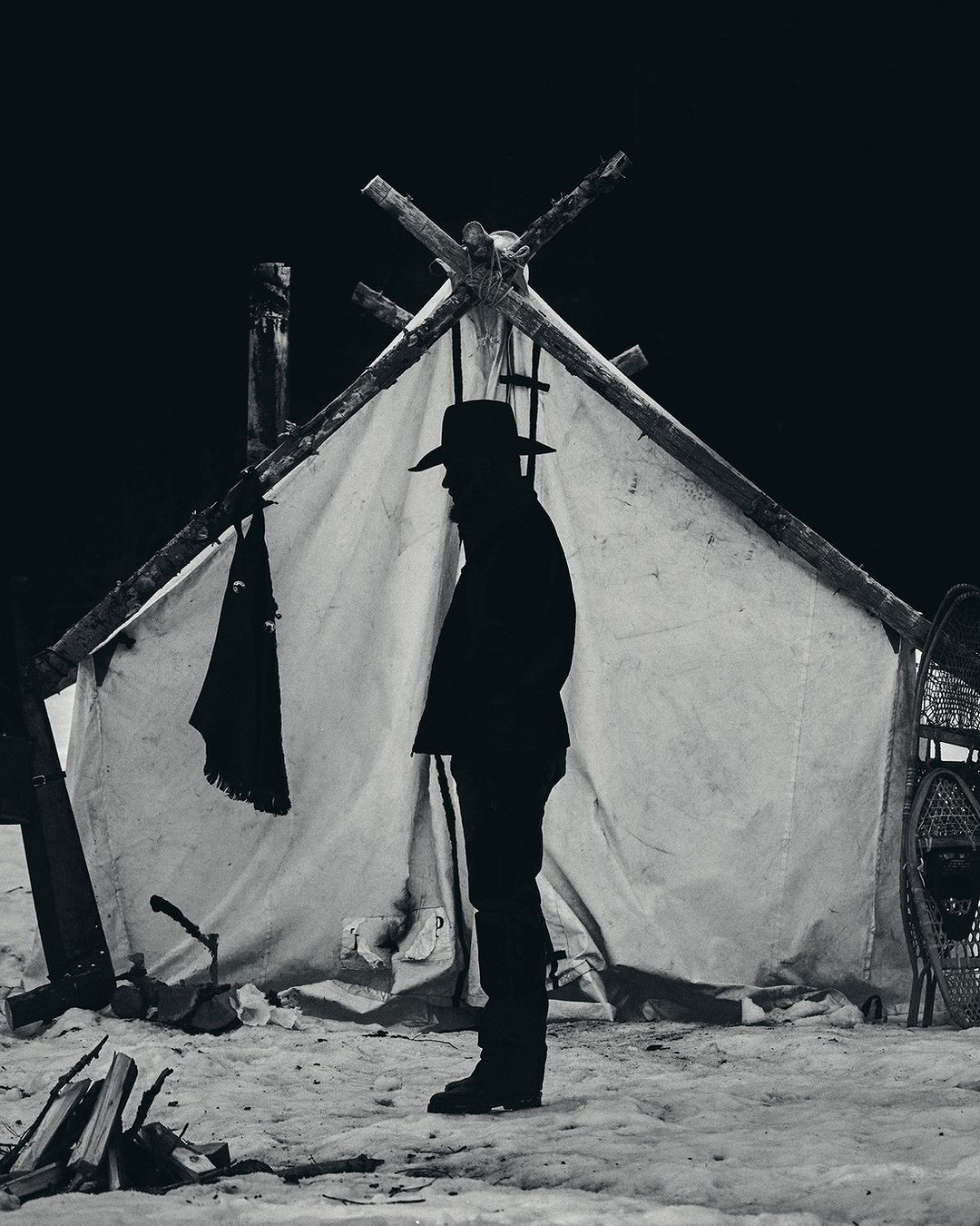 Monochrome image of a silhouetted man in a hat standing outside a canvas tent in a snowy landscape, with snowshoes and a woodpile visible.
