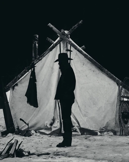 Monochrome image of a silhouetted man in a hat standing outside a canvas tent in a snowy landscape, with snowshoes and a woodpile visible.