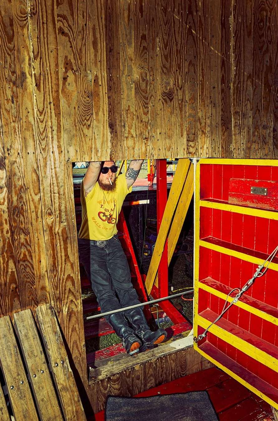 Adult male hanging from wooden tower with 'I'm A Real Cycle Slut' t-shirt, dark jeans, and sunglasses. Colorful tower interior, indoor shot.