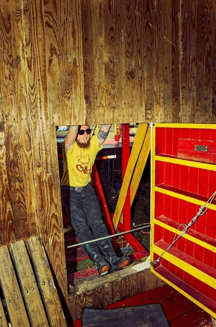 Adult male hanging from wooden tower with 'I'm A Real Cycle Slut' t-shirt, dark jeans, and sunglasses. Colorful tower interior, indoor shot.