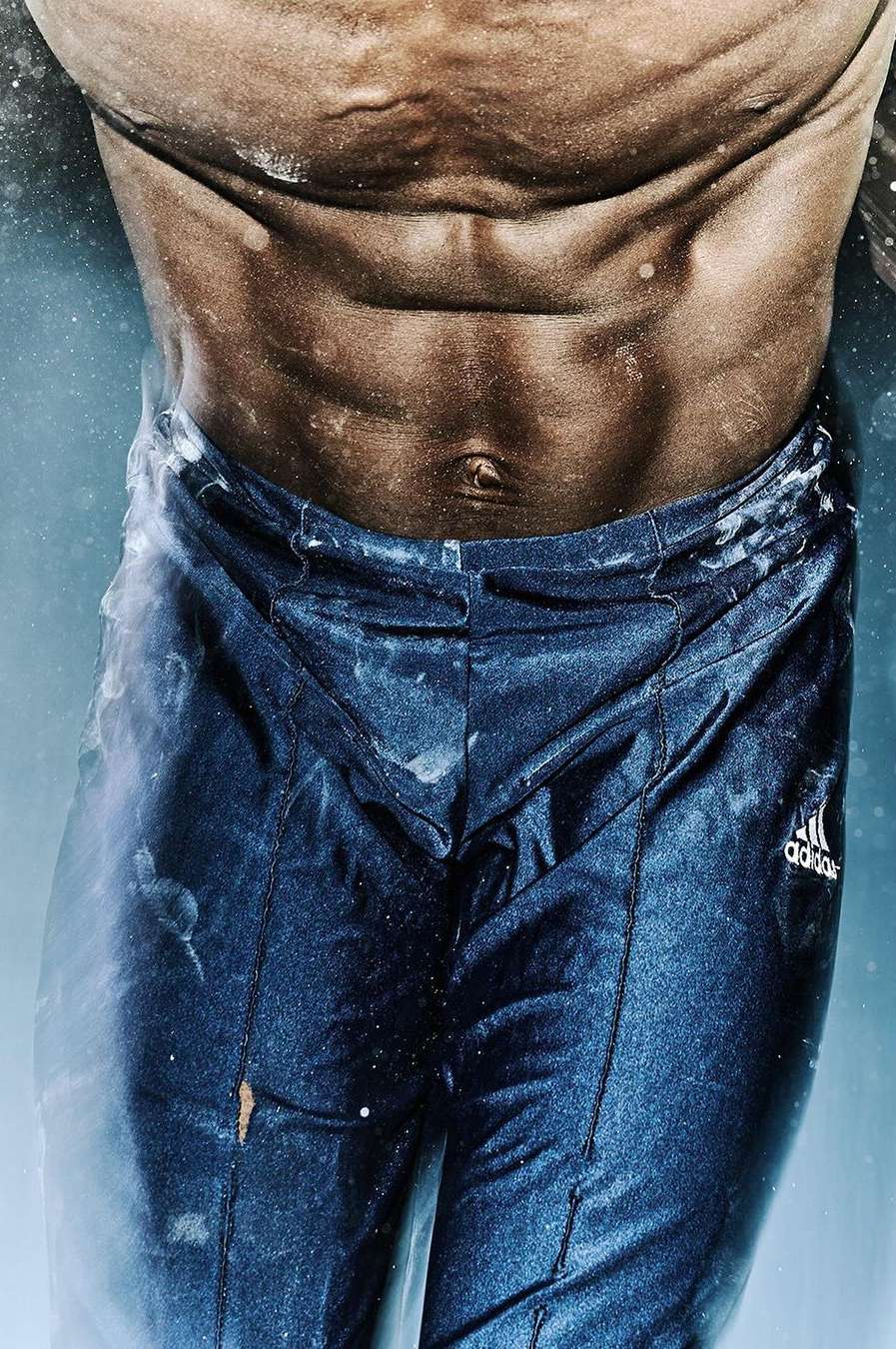 Close-up of the torso of a muscular man, showing his abs and waist with blue athletic pants, dust particles in the air.