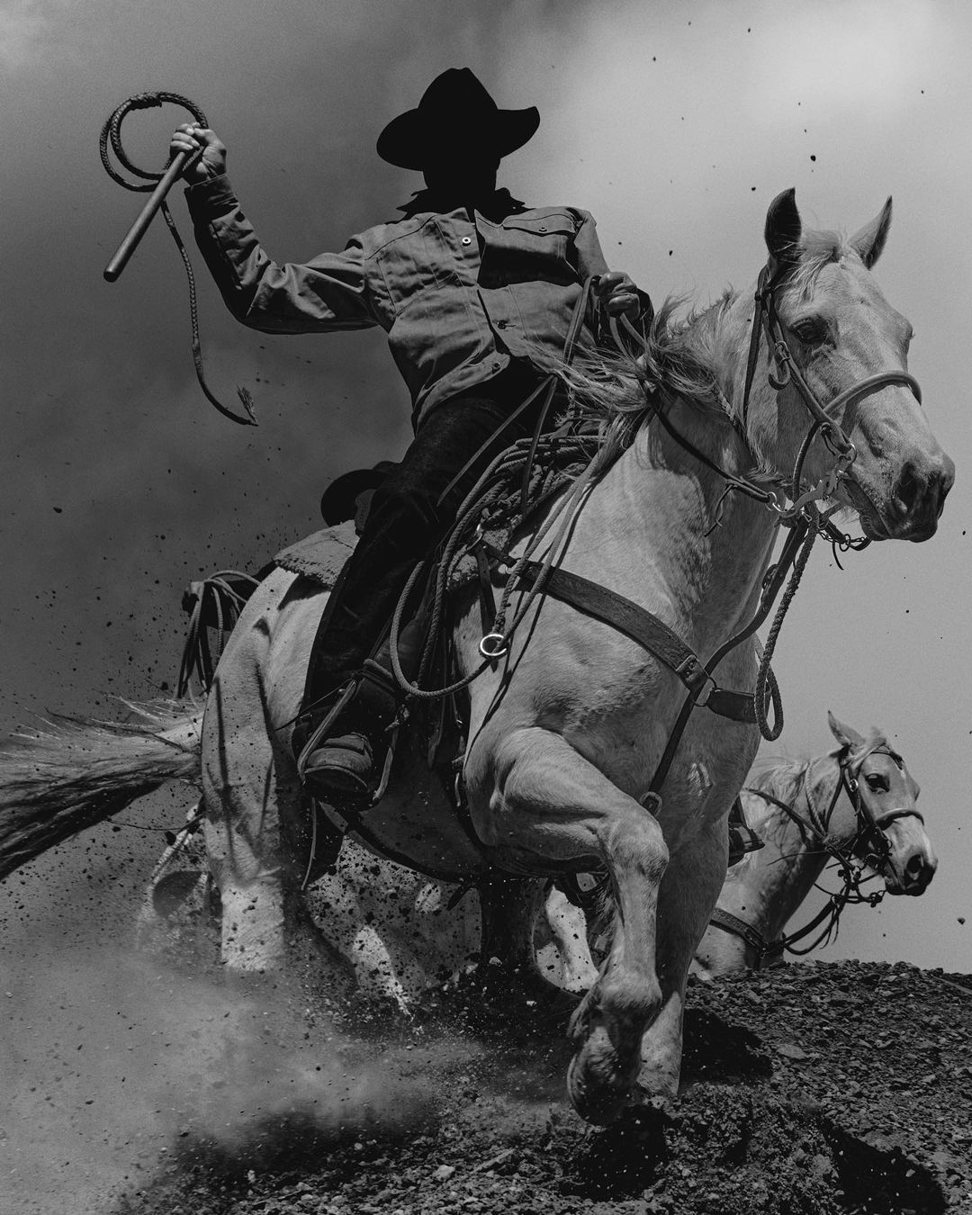 Black and white photo of a cowboy riding a horse, wielding a whip, with a second horse visible in the background.