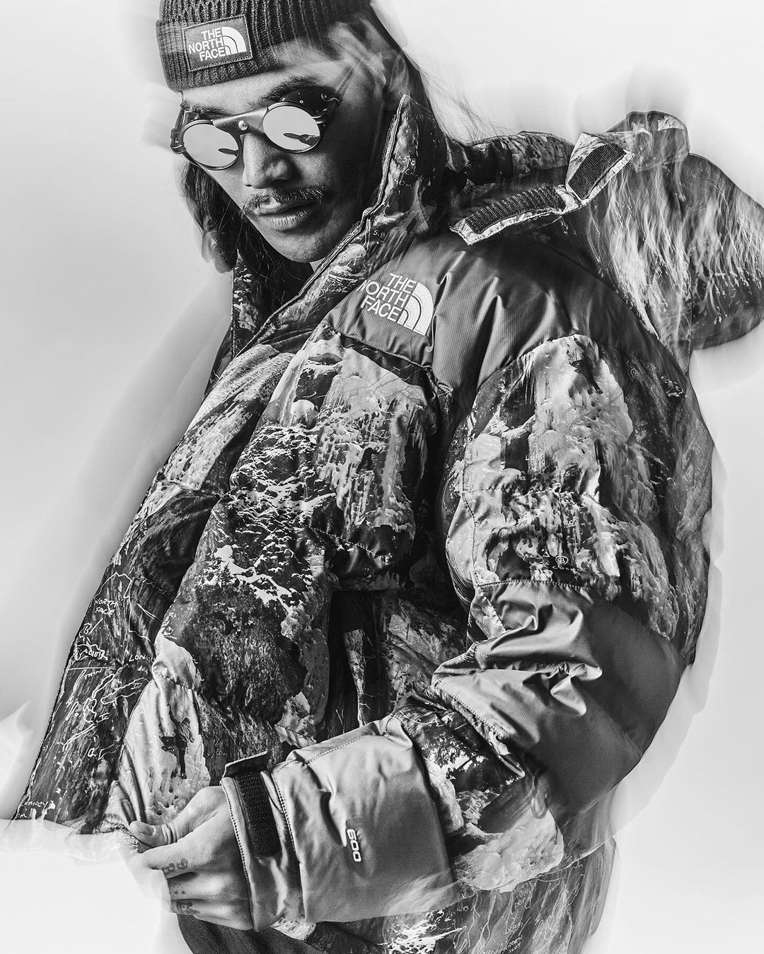 Man wearing patterned puffer jacket and beanie, posing indoors in monochrome, eyewear, and a mustache visible, lit by studio lighting.