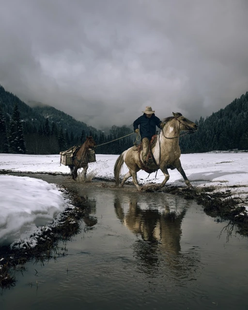 A cowboy rides a horse through shallow water with a packhorse in tow, in a snowy field surrounded by mountains and forest under a cloudy sky.