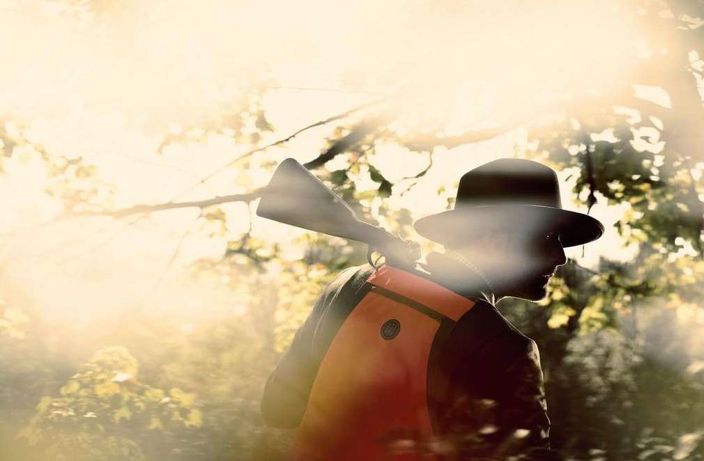 Hunter in a forest wearing a wide-brimmed hat and an orange vest, carrying a shotgun over his shoulder in a sunlit, wooded area.