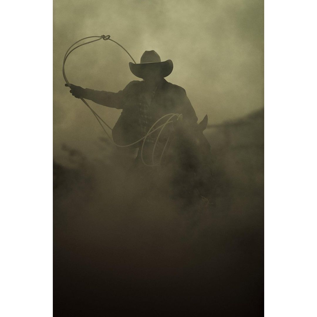A cowboy rides a horse in a sepia-toned image, holding a lasso, showcasing a Western theme in a smoky, dusty outdoor setting.