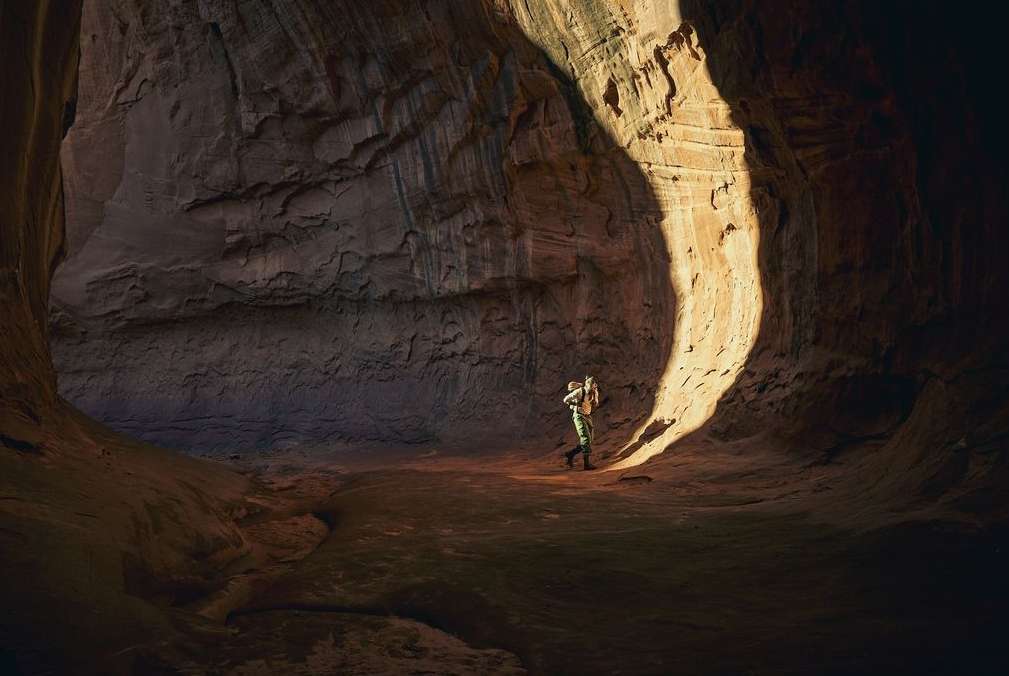 A lone hiker explores a sandstone slot canyon, lit by a beam of sunlight filtering through the narrow opening above, creating dramatic shadows.