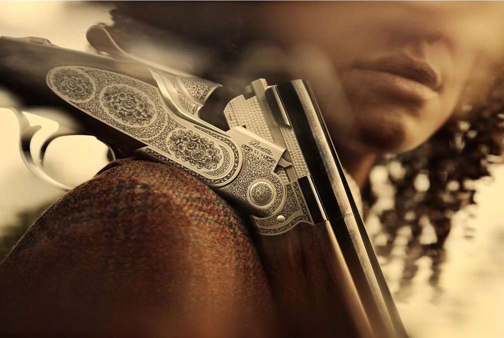Close-up of an ornately engraved Beretta shotgun lying on a person's shoulder, emphasizing the gun's details and design against a blurred background.