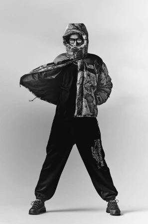 Model in hooded puffer jacket and sunglasses opening the coat against a plain background in a black and white portrait.
