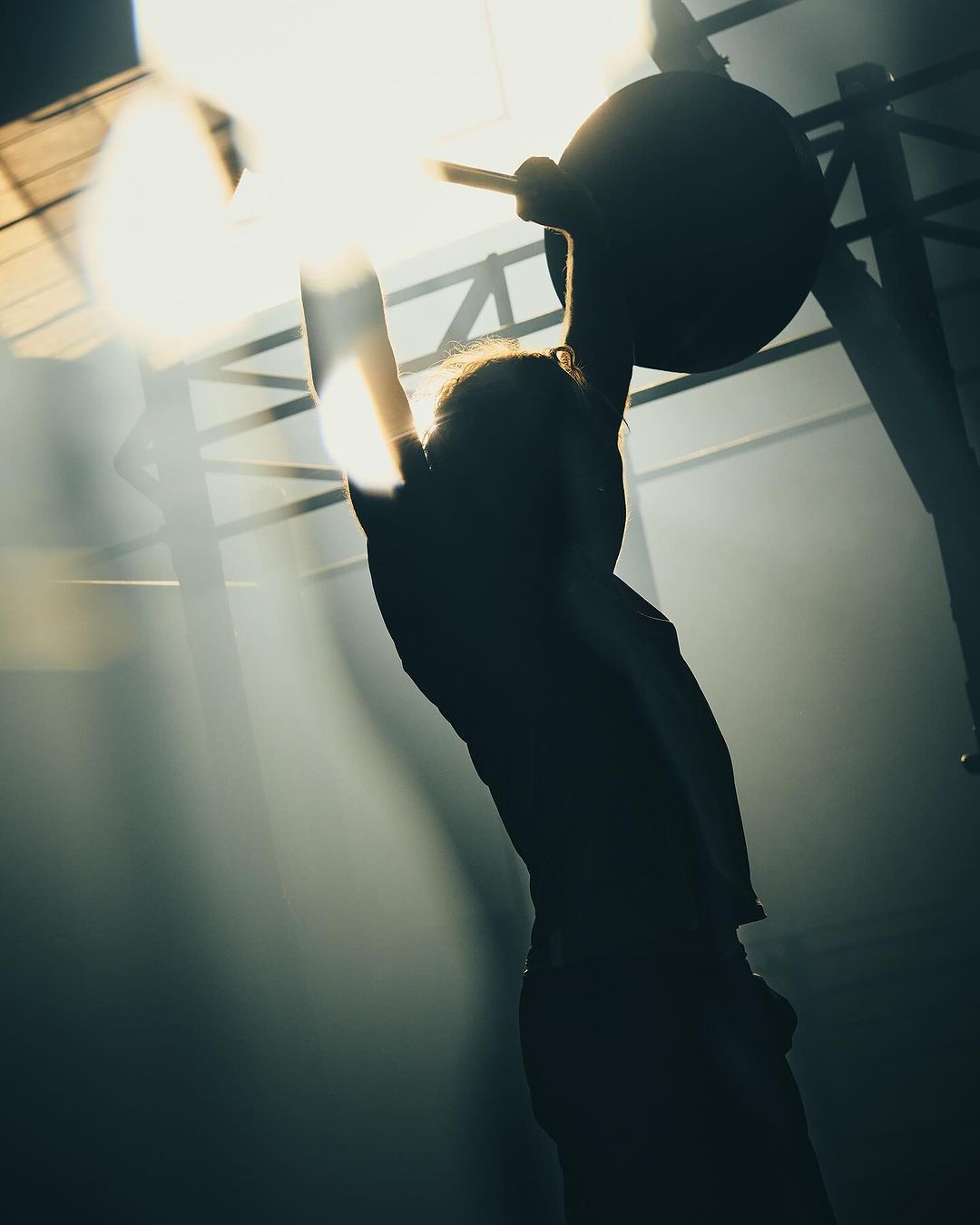 Silhouette of an adult man lifting a barbell in a dimly lit gym with bright light and sun rays creating a dramatic, high contrast effect.