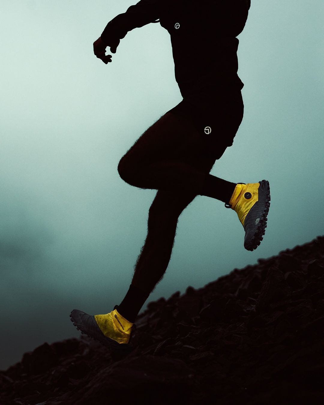 Athlete mid-motion on a hillside, athletic yellow shoes highlighted, silhouetted figure against a muted sky, dynamic pose.
