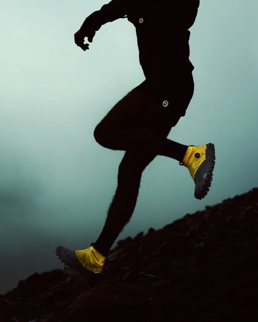 Athlete mid-motion on a hillside, athletic yellow shoes highlighted, silhouetted figure against a muted sky, dynamic pose.