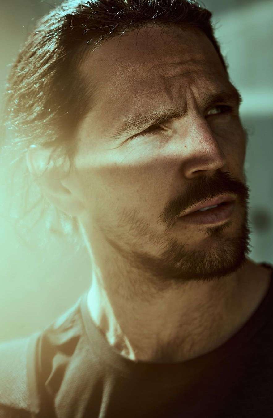 Close-up studio shot of an adult male with dark hair and a beard looking up and to the side with neutral expression and black shirt.