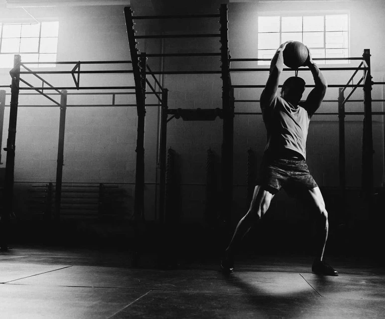 Black and white shot of an adult man in a gym, holding a medicine ball above his head while exercising near workout equipment.