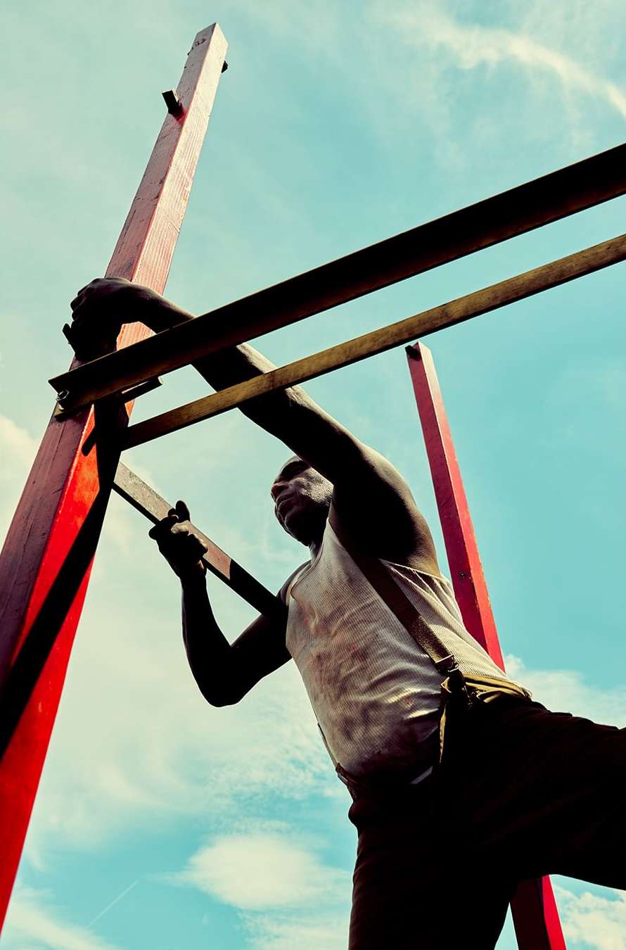 Low angle view of a Black adult male climbing a wooden structure against a blue sky with soft clouds. The man is wearing a white top.