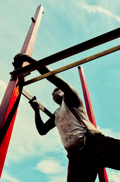 Low angle view of a Black adult male climbing a wooden structure against a blue sky with soft clouds. The man is wearing a white top.