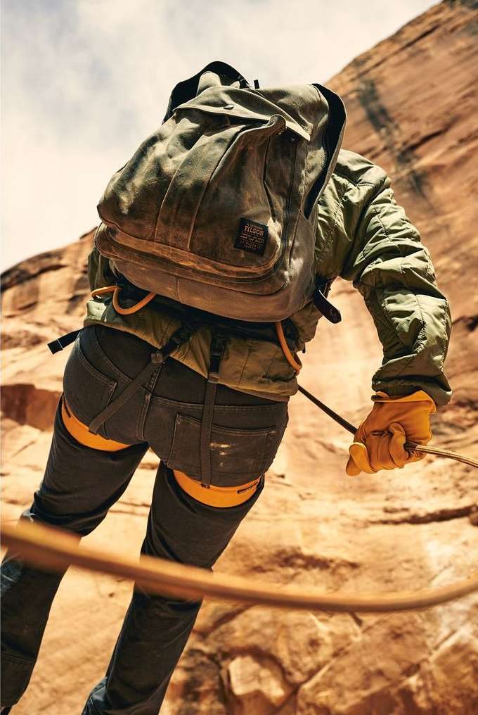 Person rappelling down a rock face with a backpack, wearing gloves and harness, under a partly cloudy sky. Outdoor adventure.