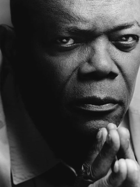 Monochrome portrait of an adult man with dark skin, looking intently at the camera with his chin resting on his clasped hands.