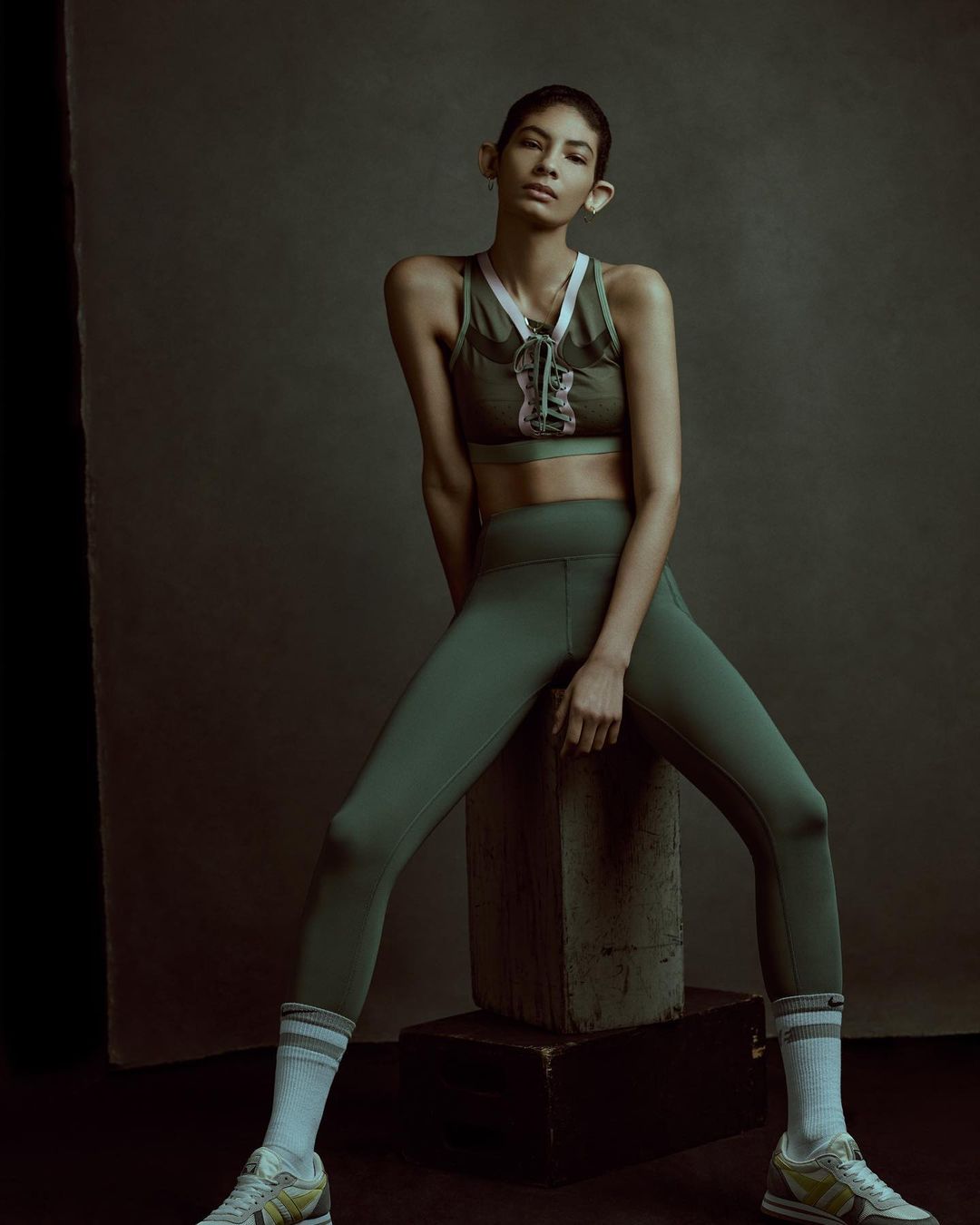 Athletic woman poses in green workout gear, sitting on a gray block in a studio setting, showcasing fashion and fitness.