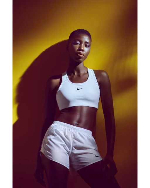 Athletic Black woman poses in white sports bra and shorts in front of yellow wall with shadow, fitness apparel.
