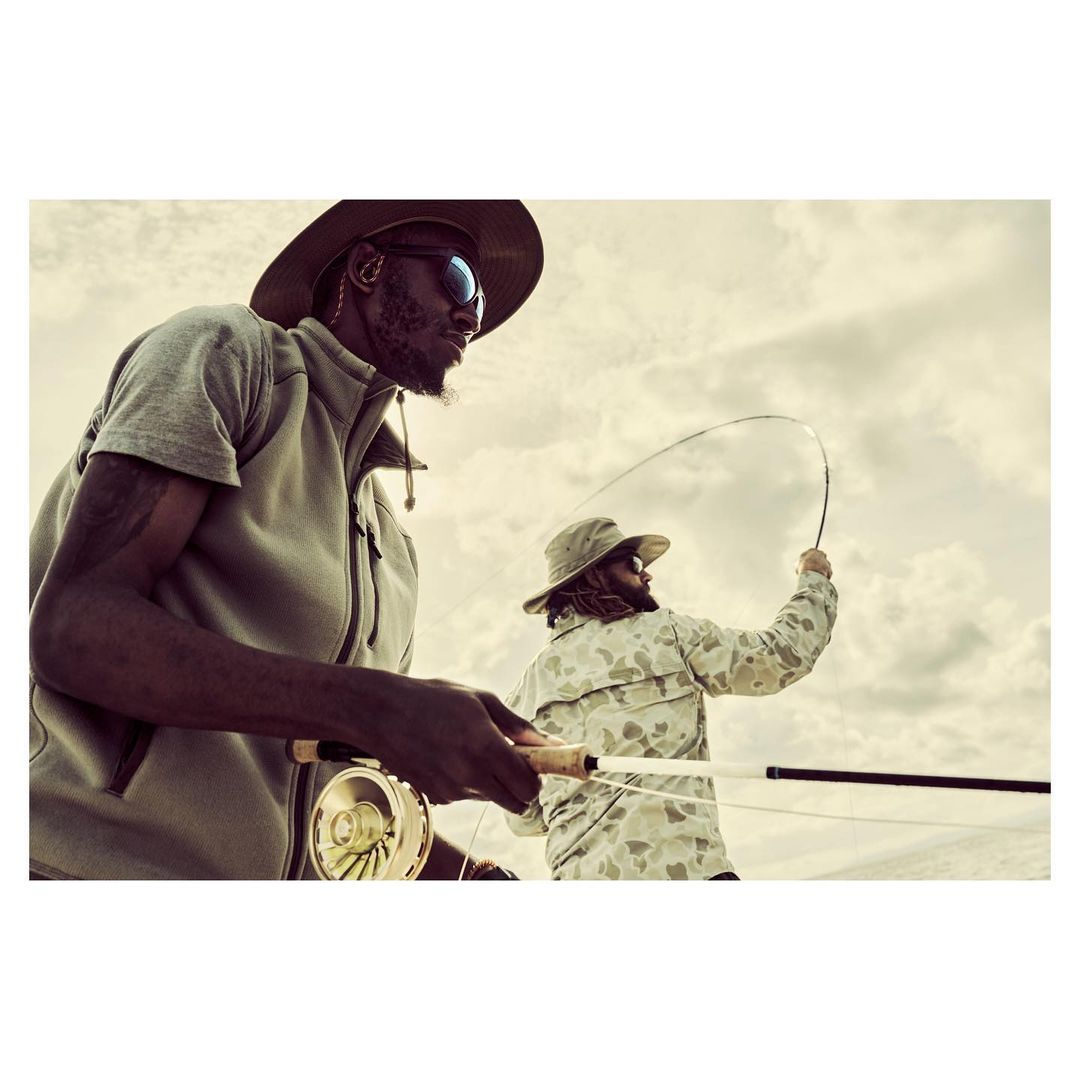 Two men fly fishing under a cloudy sky. The man in the foreground is wearing sunglasses and a hat. He is holding a fishing rod.