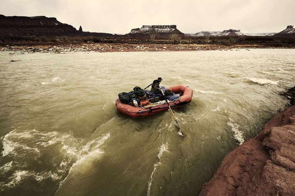 An adult rows a red inflatable raft loaded with gear on a flowing river with arid rock formations and cliffs in the background on an overcast day.