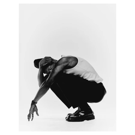 A Black man wearing a white tank top and dark trousers crouches low against a seamless white background in New York.