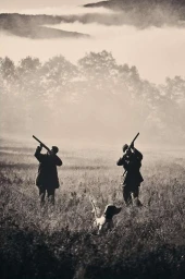 Two figures holding hunting rifles in a misty field with a dog in the foreground captured in a sepia-toned editorial photograph.